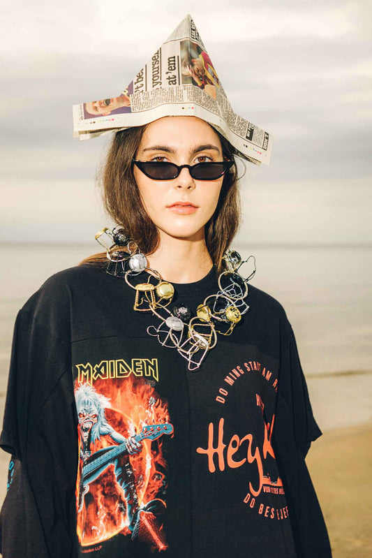 Close-up of model wearing a black oversized dress with graphic designs and text, accessorized with a newspaper hat and cork muselet necklace.